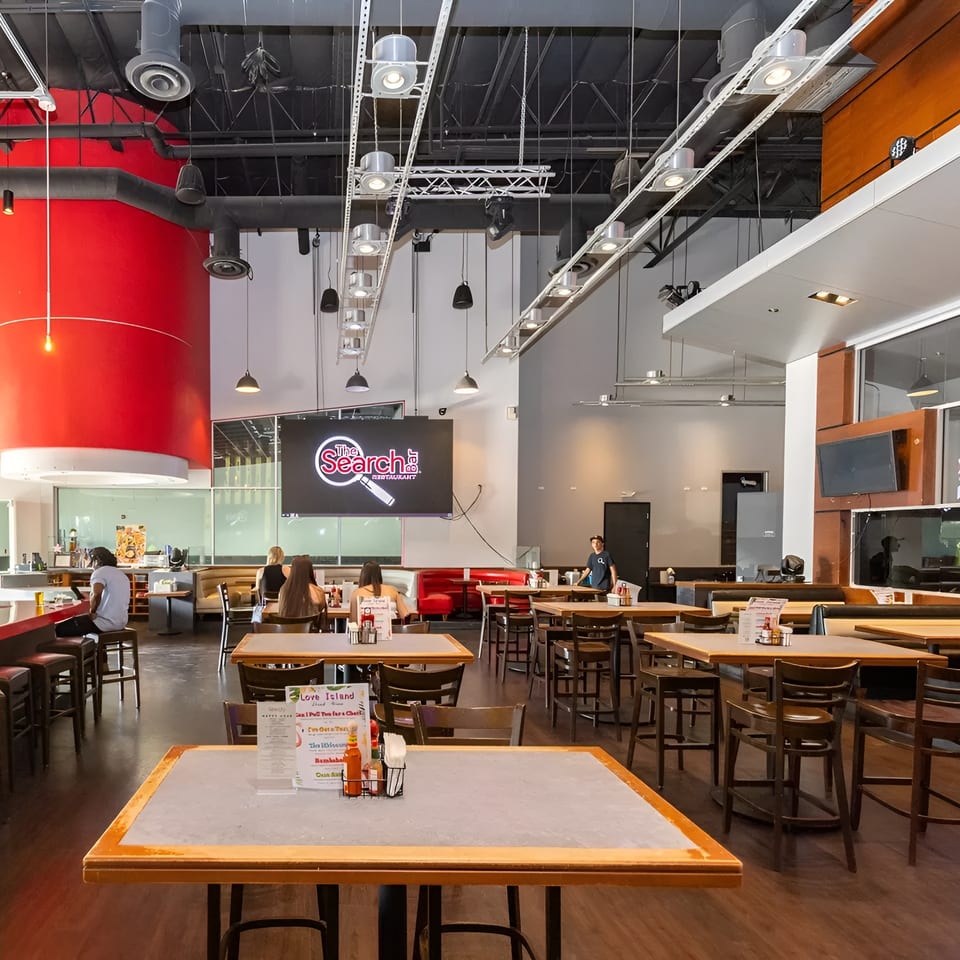 A spacious restaurant interior with high ceilings, a red bar, and multiple dining tables with patrons.