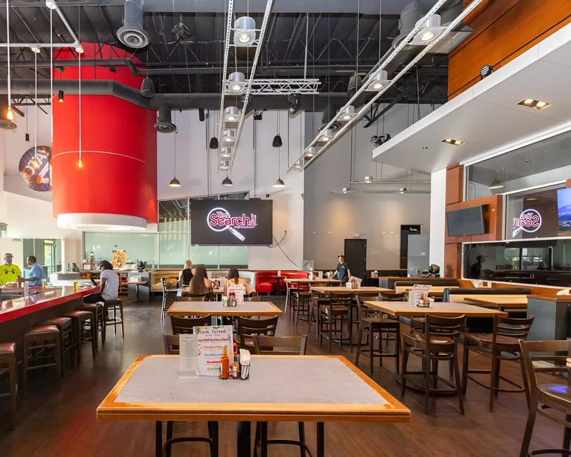 A spacious restaurant interior with high ceilings, a red bar, and multiple dining tables with patrons.