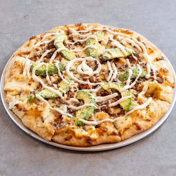 A whole pizza topped with cheese, bacon, avocado slices, and a white sauce drizzle on a metal pan.