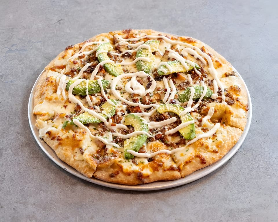 A whole pizza topped with cheese, bacon, avocado slices, and a white sauce drizzle on a metal pan.
