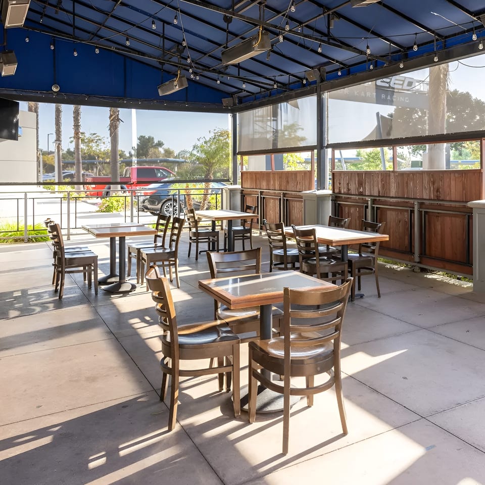 An empty outdoor patio with wooden tables and chairs under a blue awning on a sunny day.