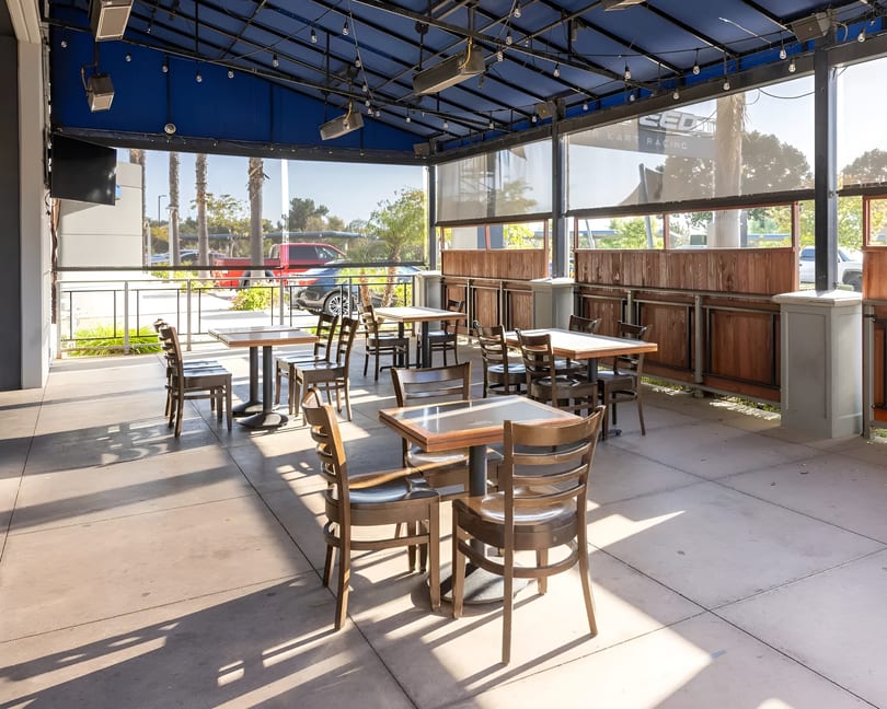 An empty outdoor patio with wooden tables and chairs under a blue awning on a sunny day.
