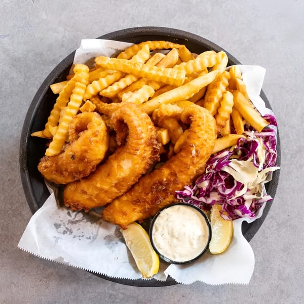 A black basket of golden fish and chips with coleslaw, tartar sauce, and lemon wedges.