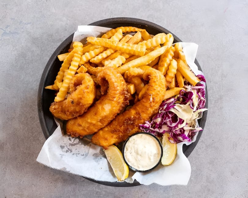 A black basket of golden fish and chips with coleslaw, tartar sauce, and lemon wedges.