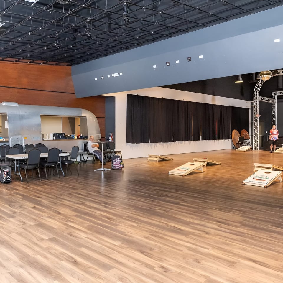 Spacious event hall with a bar, tables, cornhole games, and people socializing.