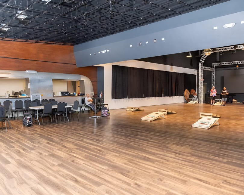 Spacious event hall with a bar, tables, cornhole games, and people socializing.