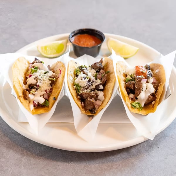Three street tacos with meat, cheese, and sauce, served with lime wedges and salsa.