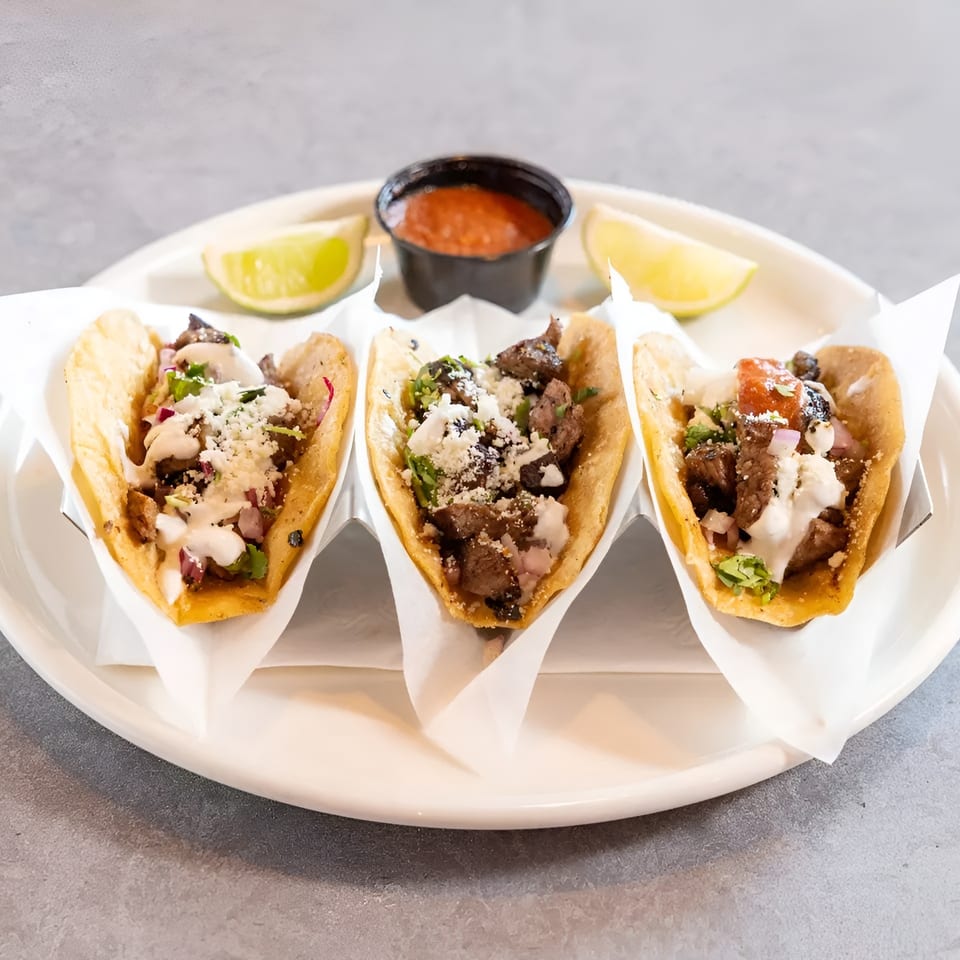 Three street tacos with meat, cheese, and sauce, served with lime wedges and salsa.