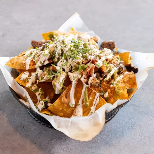 A basket of loaded nachos with meat, cheese, sauces, and cilantro on a gray surface.