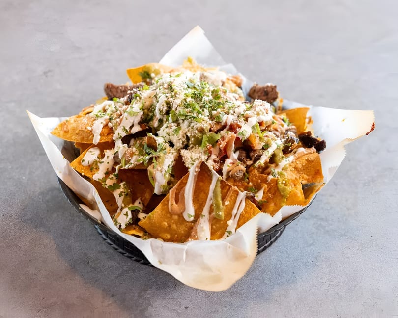 A basket of loaded nachos with meat, cheese, sauces, and cilantro on a gray surface.