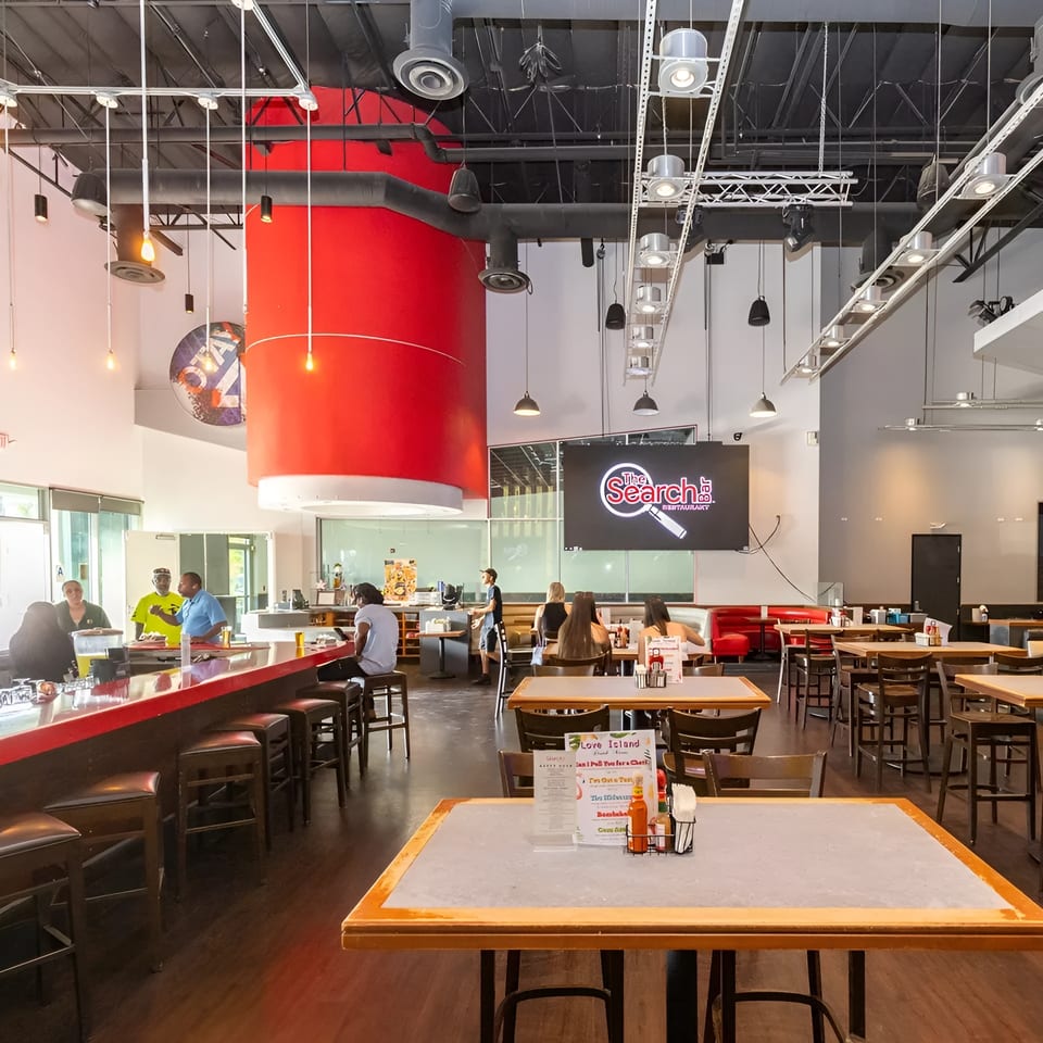 Spacious restaurant interior with a red bar, dining tables, and a large screen displaying "The Search Restaurant" logo.