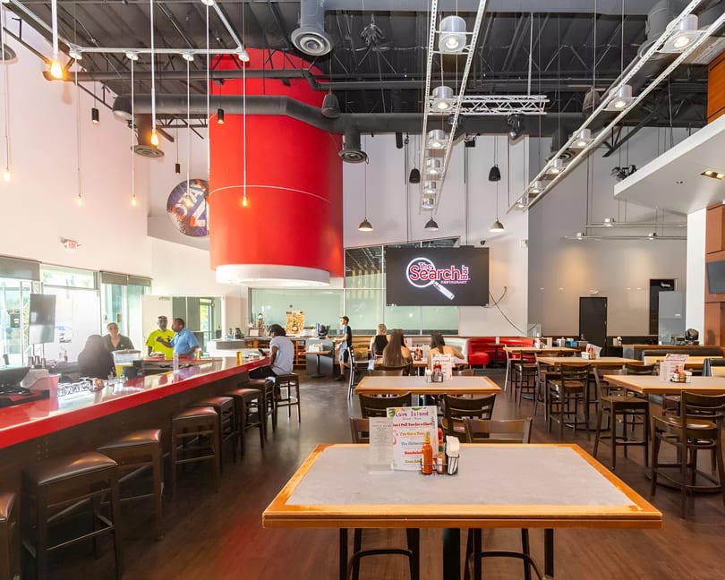 Spacious restaurant interior with a red bar, dining tables, and a large screen displaying "The Search Restaurant" logo.