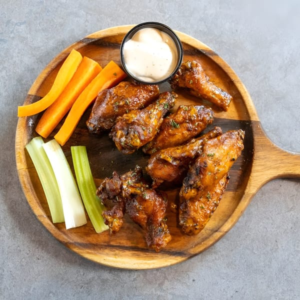 Glazed chicken wings with carrots, celery, and dipping sauce on a wooden serving board.