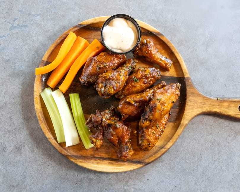 Glazed chicken wings with carrots, celery, and dipping sauce on a wooden serving board.