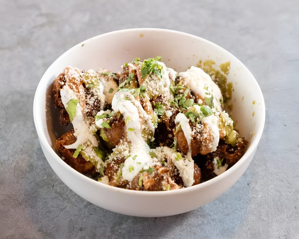 A white bowl filled with crispy fried brussels sprouts, drizzled with white sauce, green sauce, and cilantro.