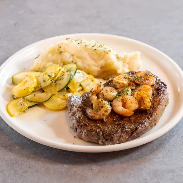 Steak and shrimp with mashed potatoes and sautéed zucchini and squash on a white plate.