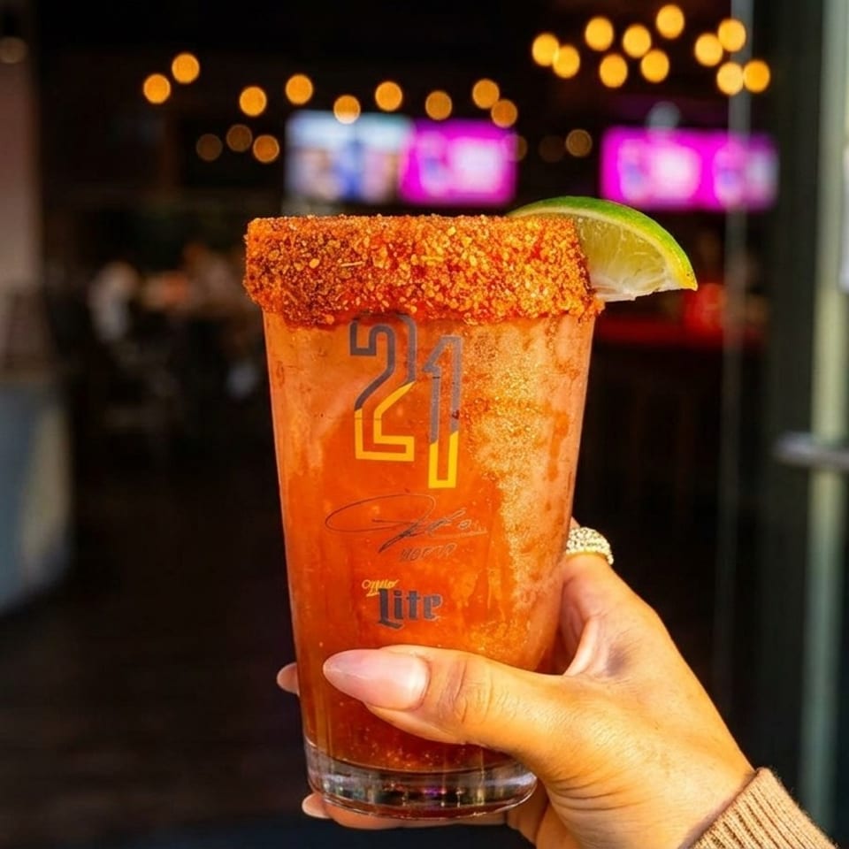 A hand holds a michelada with a chili-lime rim and lime wedge, against a blurred restaurant background.