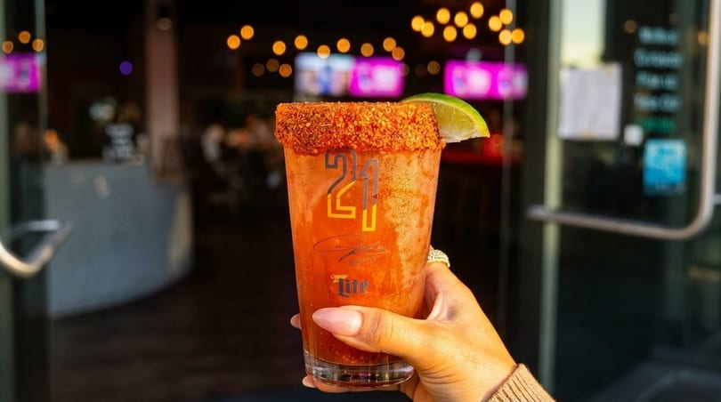 A hand holds a michelada with a chili-lime rim and lime wedge, against a blurred restaurant background.