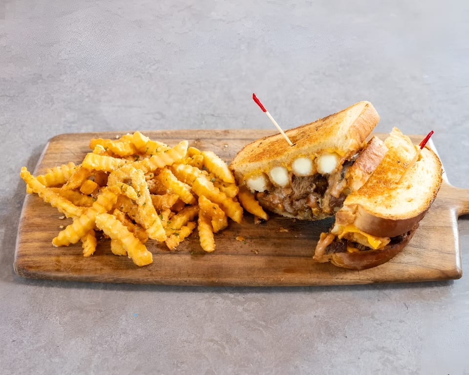 Grilled cheese sandwich with meat and cheese curds, served with crinkle-cut fries on a wooden board.