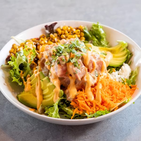 A colorful poke bowl with tuna, avocado, corn, shredded carrots, and greens, drizzled with sauce.