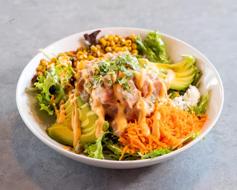A colorful poke bowl with tuna, avocado, corn, shredded carrots, and greens, drizzled with sauce.