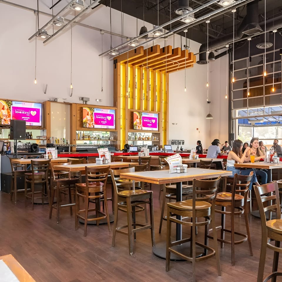 A spacious restaurant interior with high-top tables, bar seating, and large screens displaying "Love Island."