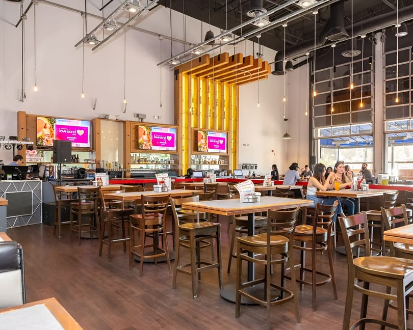 A spacious restaurant interior with high-top tables, bar seating, and large screens displaying "Love Island."
