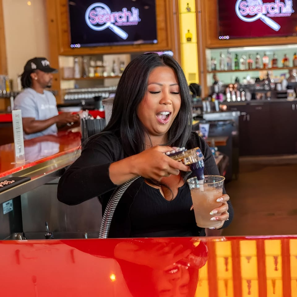 A female bartender pours a drink at a red bar, with another bartender in the background.
