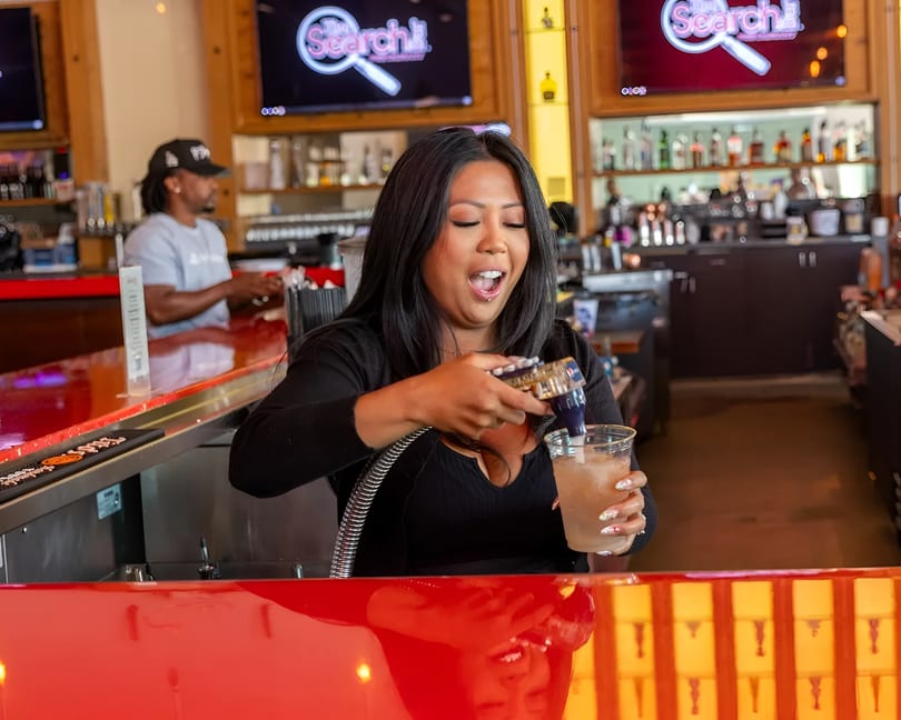 A female bartender pours a drink at a red bar, with another bartender in the background.
