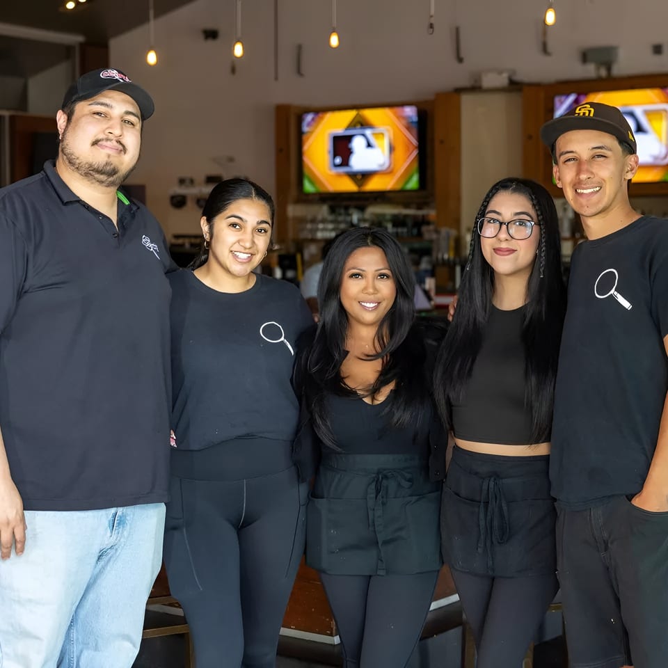Five smiling restaurant staff members in black uniforms stand together in a casual dining area.