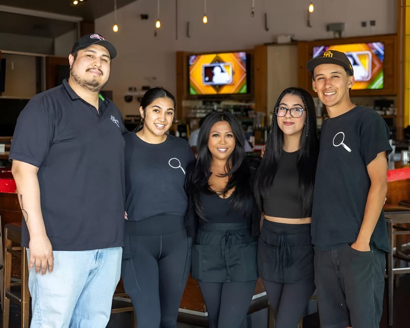 Five smiling restaurant staff members in black uniforms stand together in a casual dining area.