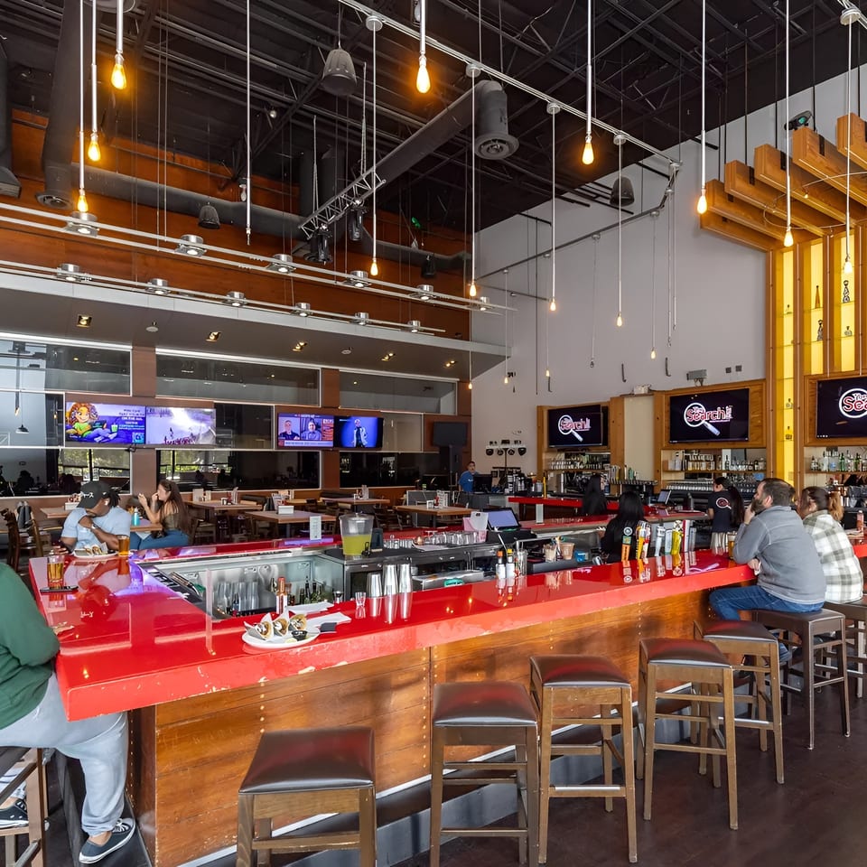 A vibrant, modern restaurant interior with a long red bar, patrons, and large TV screens.