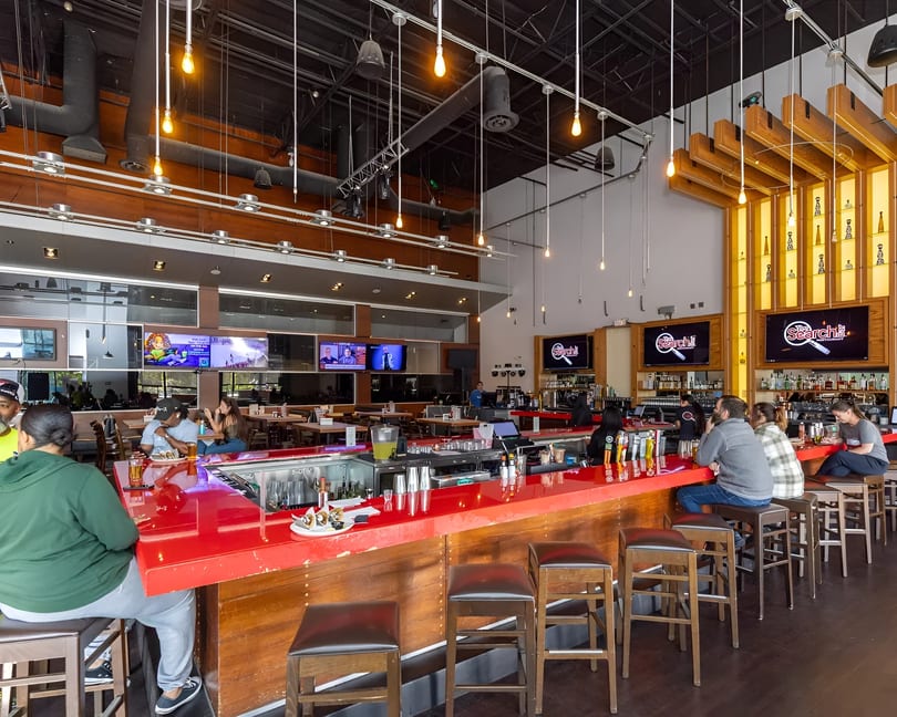 A vibrant, modern restaurant interior with a long red bar, patrons, and large TV screens.
