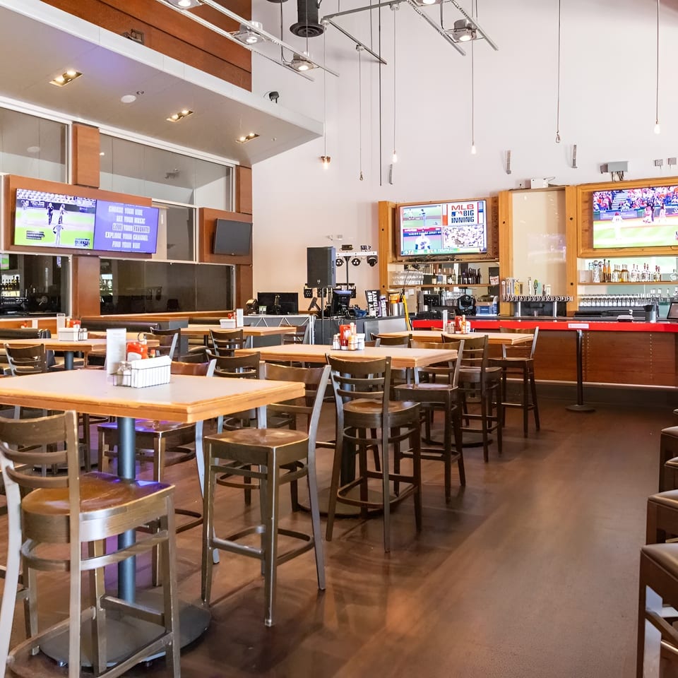 Spacious sports bar interior with high-top tables, stools, a long red bar, and multiple TVs showing baseball.