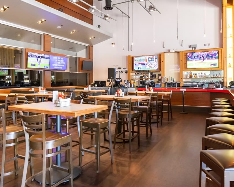 Spacious sports bar interior with high-top tables, stools, a long red bar, and multiple TVs showing baseball.